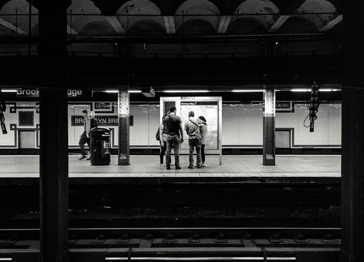 Black‑and‑white photo of people standing on a subway platform near an information display.