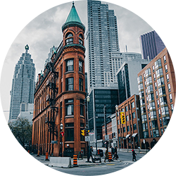 Urban street corner with a historic red-brick flatiron-style building, surrounded by modern high-rises and people crossing the street in Toronto