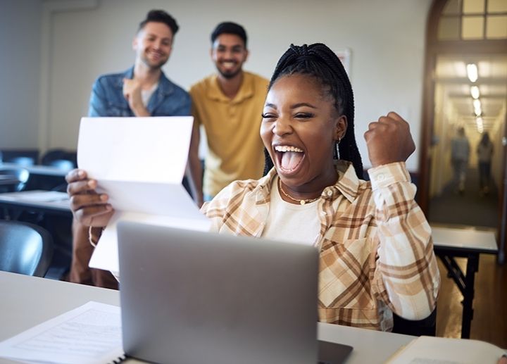 Person celebrating while reading a document at a laptop, with others standing nearby